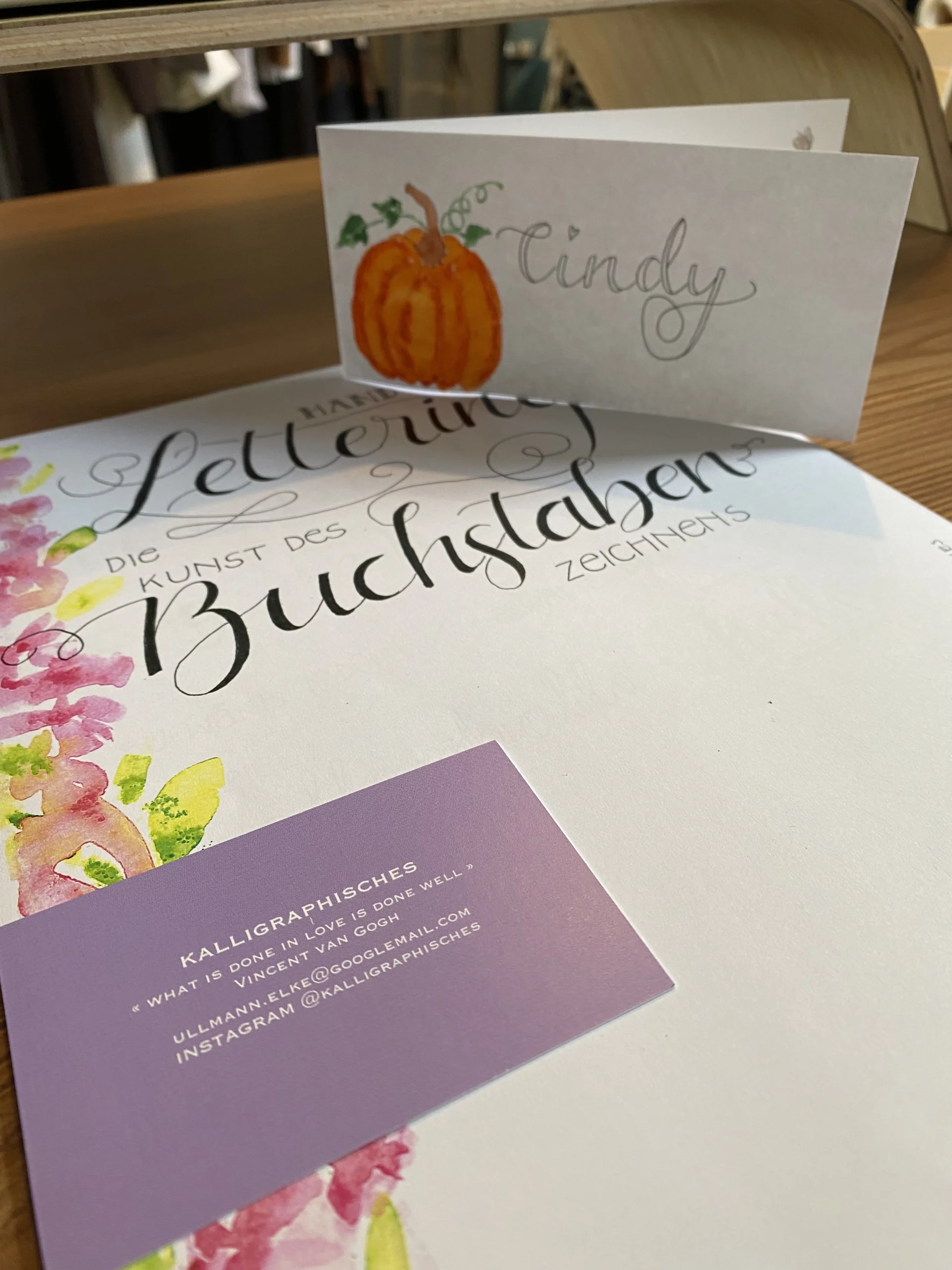 Nahaufnahme eines liebevoll gestalteten Namensschilds mit Kürbiszeichnung, einem Handlettering-Übungsbuch und einer Visitenkarte auf einem Holztisch – aufgenommen im Rahmen eines Kreativ-Workshops bei Cindys Frauenzimmer in Schneverdingen.